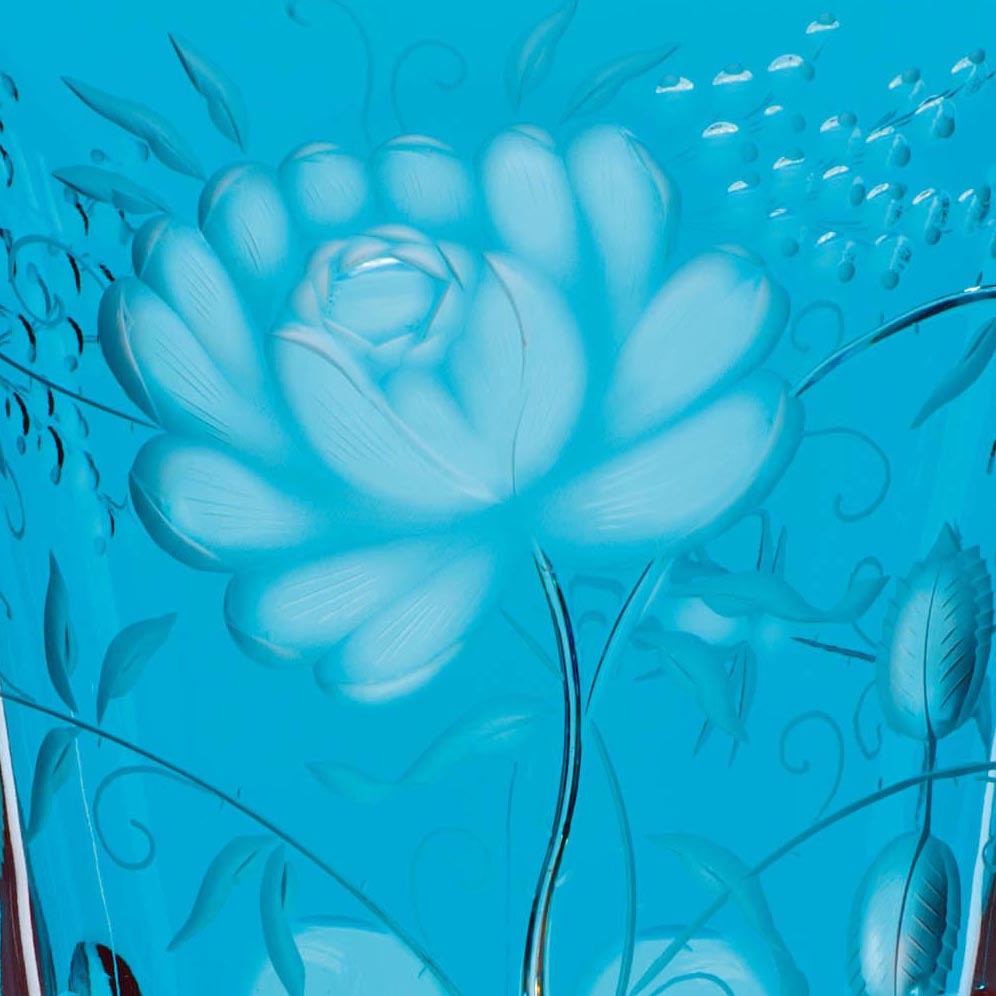 Close-up of an azure crystal vase. A large, intricately cut floral motif with many petals is engraved. Tendrils and leaves surround the flower. The brilliant cut showcases the craftsmanship.