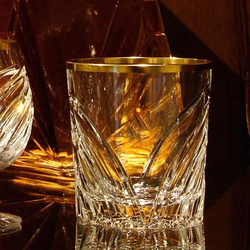 An intricately designed crystal whisky glass from the Wings series, featuring a striking gold rim and opulent, hand-cut patterns. The clear, heavy glass reflects warm, golden light in the style of the Golden Twenties.