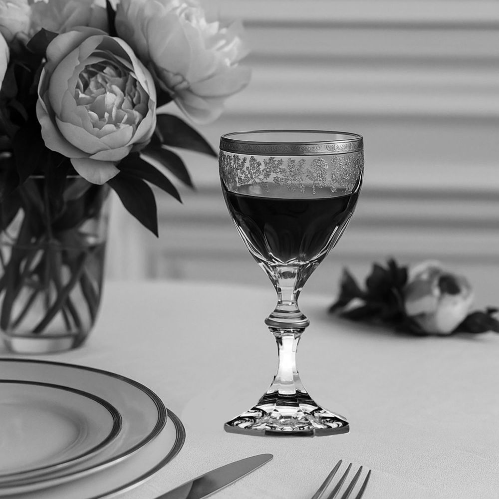 Elegant crystal red wine glass with dark wine on a light-coloured table. Rim and bowl have a floral pattern; slender stem and faceted base. Background left: bouquet in a vase; foreground: plate and cutlery.