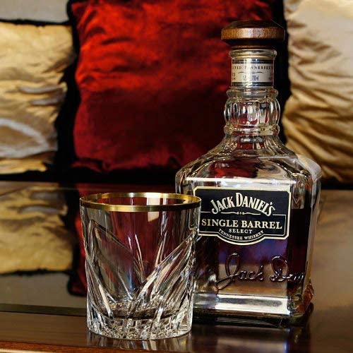 Clear crystal whisky glass by ARNSTADT KRISTALL featuring an opulent, hand-cut Wings pattern and a gold rim on a dark wooden base. Next to it, a square whisky bottle with a dark label. Out of focus: red and gold-beige cushions.