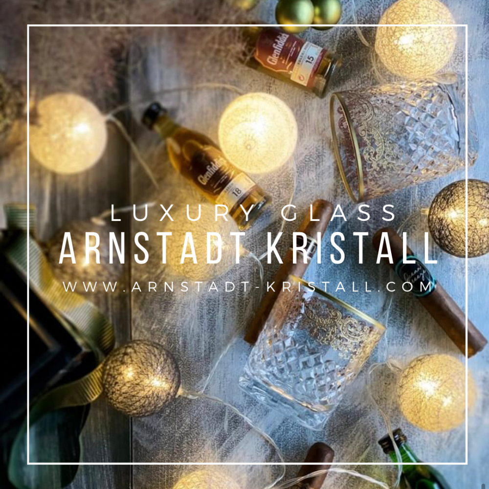 Two ‘ ARNSTADT KRISTALL ’ whisky glasses crafted from clear crystal with a golden arabesque pattern sit alongside mini whisky bottles and cigars. Warm, glowing spherical fairy lights create a luxurious, festive ambience.
