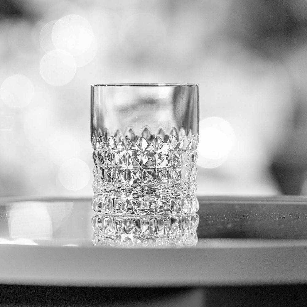 Clear crystal whisky glass in the Rocks design by ARNSTADT KRISTALL, featuring an intricately cut diamond pattern on the lower half. It stands on a reflective surface, with points of light shimmering in the blurred background.