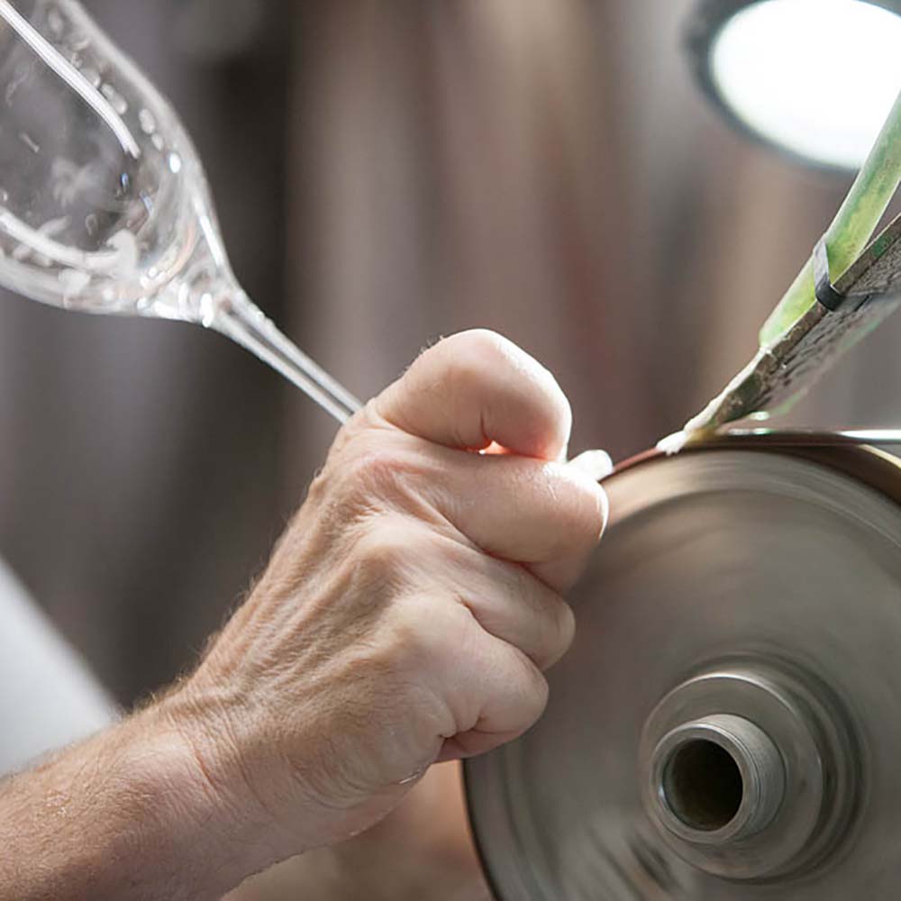 Close-up of a hand pressing a clear crystal wine glass firmly against a rapidly rotating grinding wheel to create the deep cut. Bright light in the background illuminates the process and highlights the details.