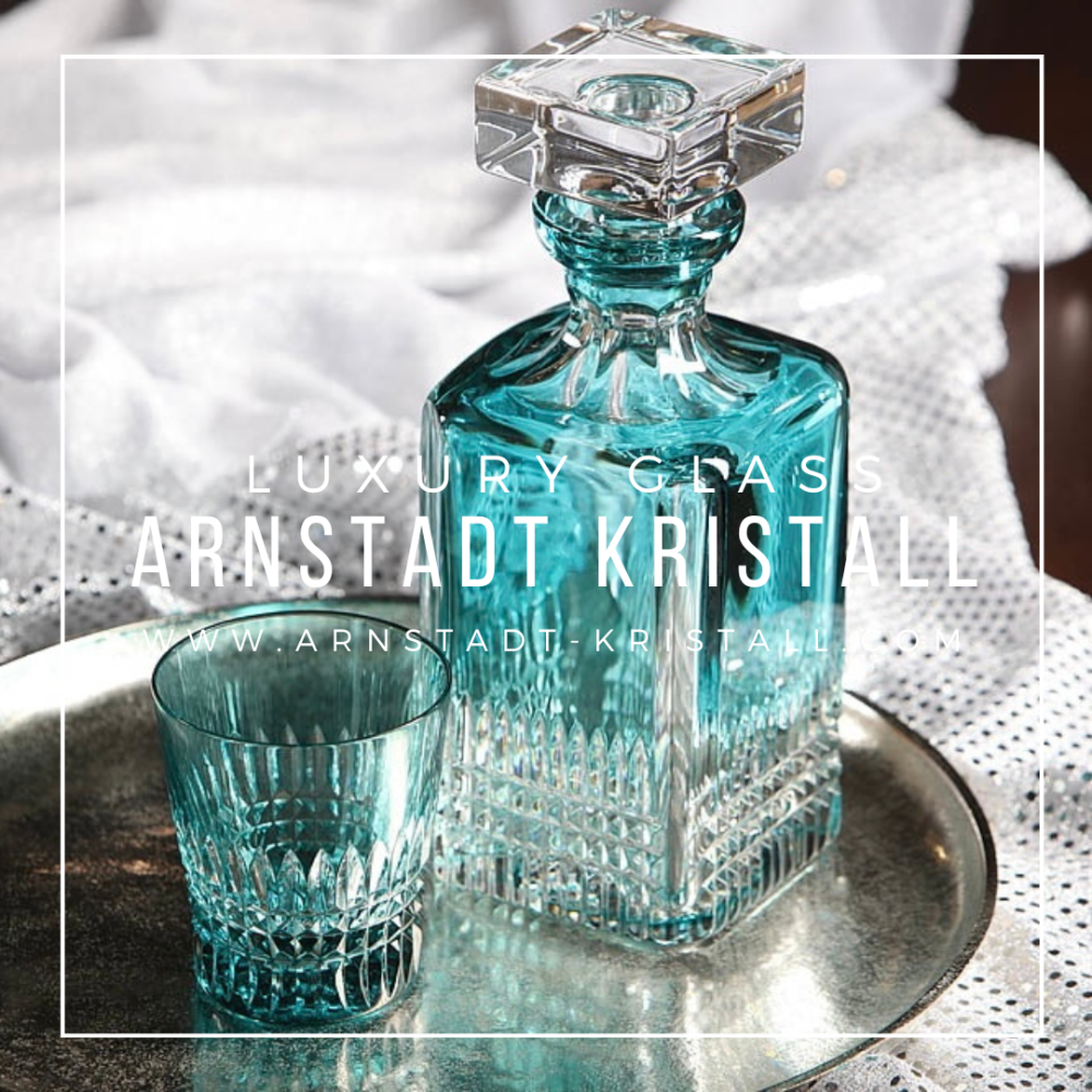 An azure blue crystal whisky decanter with a stopper and a small whisky glass sit on a round silver serving tray. Both feature sparkling faceted cuts that reflect the light. Light-coloured glittery fabric in the background.