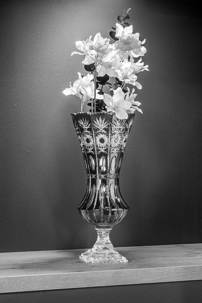 A tall, intricately cut crystal vase stands on a light-coloured wooden base. Artistic patterns and vertical grooves catch the light. Several large, light-coloured flowers with dark foliage rise elegantly from the vase.