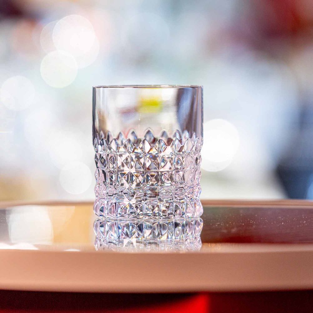 A clear cylindrical crystal whisky glass sits on a glossy surface. The lower half has a diamond-cut pattern that refracts light and conveys luxury. The upper section is smooth, while the background is soft and speckled with highlights.
