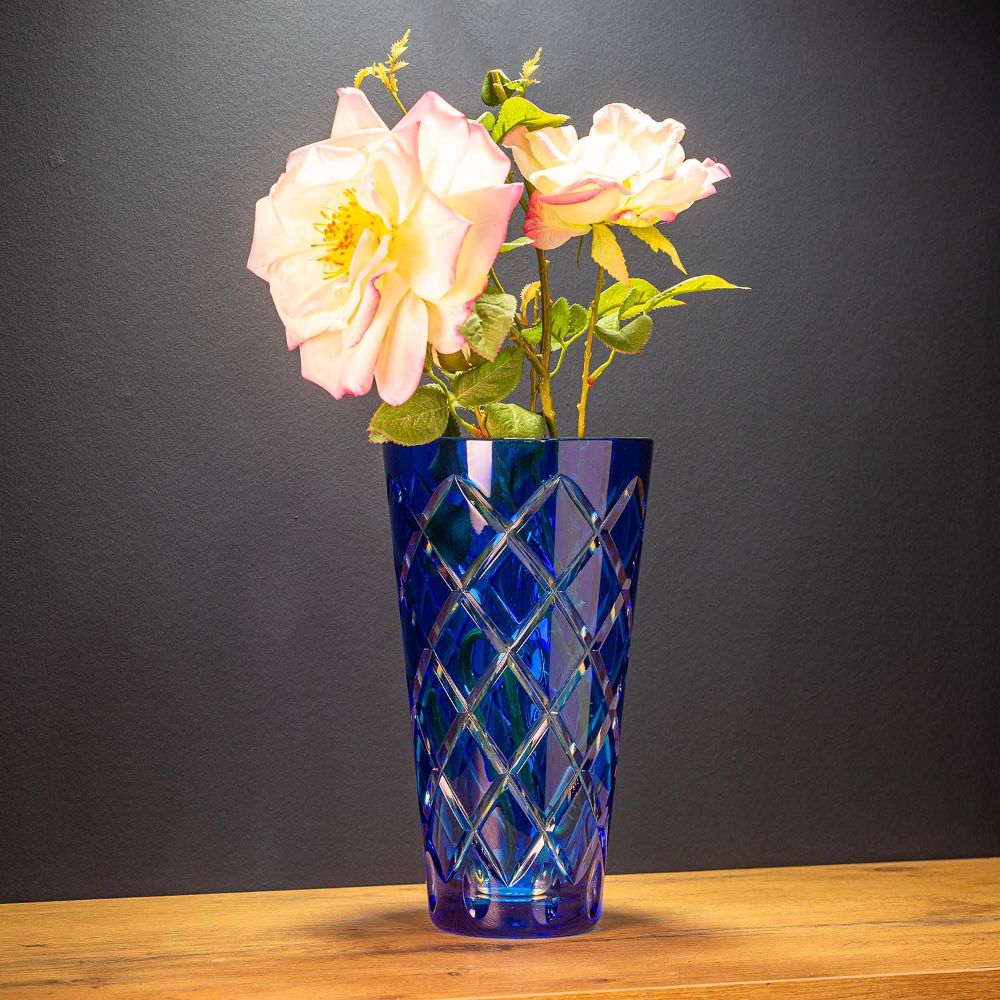 A tall, dark blue crystal vase with a brilliant diamond-cut finish, set against a light wooden surface on a dark background. The vase contains two delicate pink roses with green leaves. The cut reflects light and sparkles.