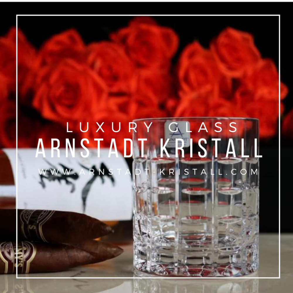 Clear crystal whisky glass, ‘Las Vegas’ model, in the foreground, featuring a geometric diamond pattern and light-refracting liquid. Blurred in the background: red roses, a bottle and two cigars, creating a luxurious atmosphere.