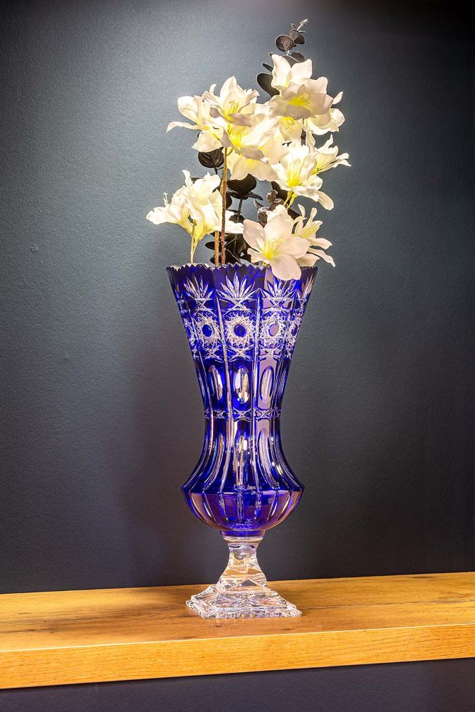 A tall, elegantly cut crystal vase in a deep cobalt blue, filled with light, creamy-white flowers and dark leaves. Set on a light brown wooden shelf against a dark wall; symmetrical cut patterns reflect the light.