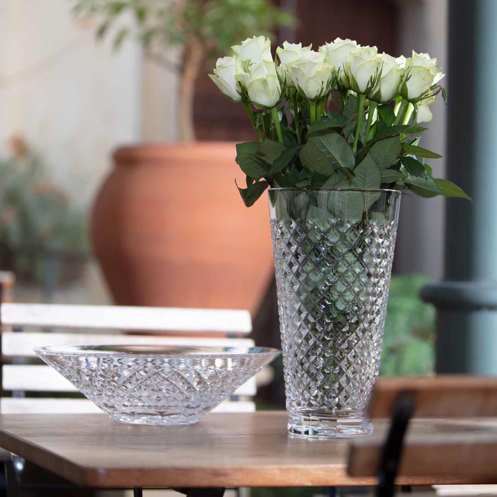 Tall ‘ ARNSTADT KRISTALL ’ vase with a diamond-cut finish, filled with white roses. Next to it, a shallow crystal bowl on a wooden table outdoors, with plants and a terracotta pot in the background.