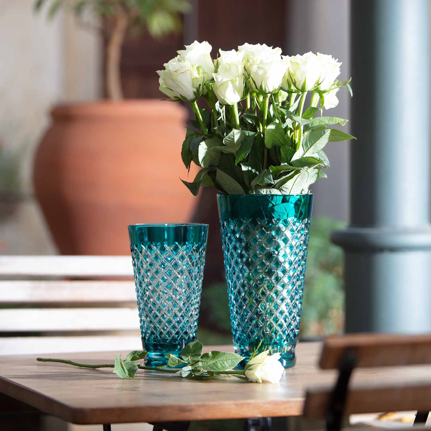 Two elegant crystal vases in azure turquoise with a chequered pattern stand on a wooden table. The larger vase contains a bouquet of white roses, with a single white rose lying next to it. In the blurred background: a terracotta pot and green plants.