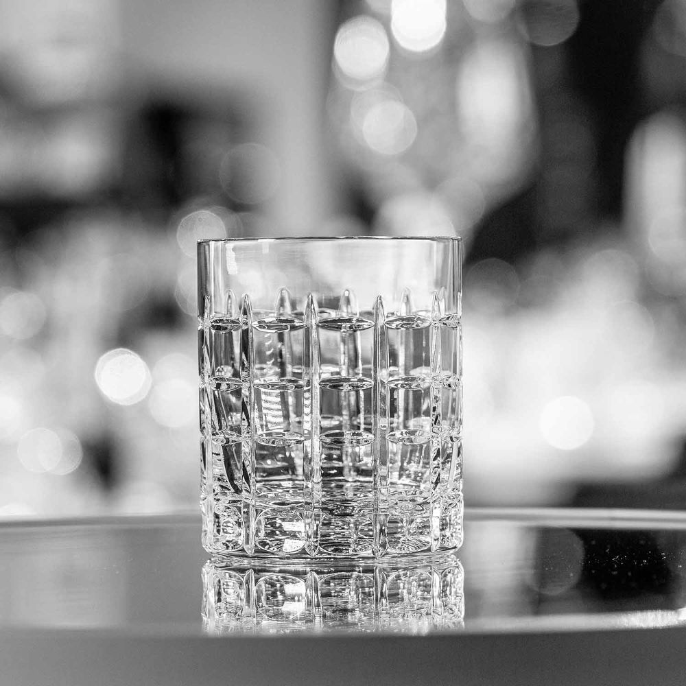 Clear, cylindrical “ ARNSTADT KRISTALL ” Las Vegas whisky glass crafted from high-quality crystal glass. Detailed, lattice-like cut pattern with small square facets on a reflective surface against a blurred background with light reflections.