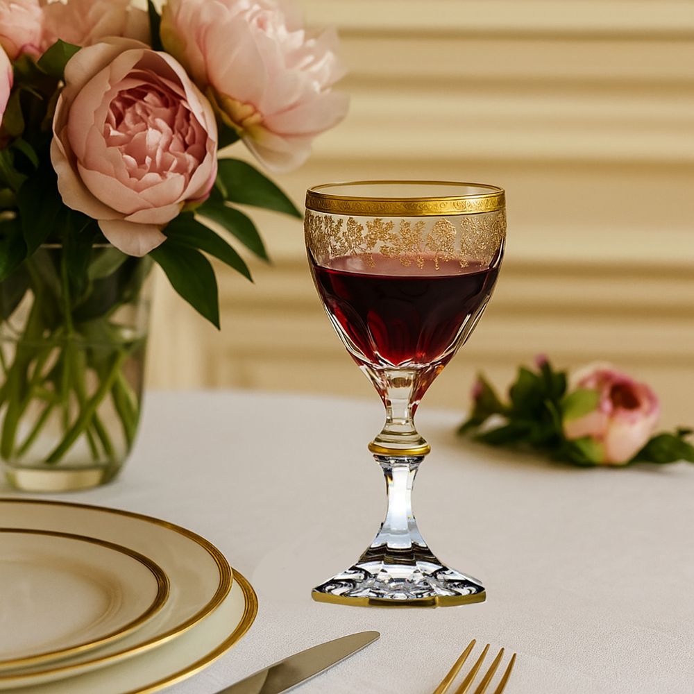 An elegant crystal red wine glass with red wine on a white table. Delicate gold ornamentation on the rim and stem. In the background, pink peonies in a vase and a festively laid table with plates and cutlery.