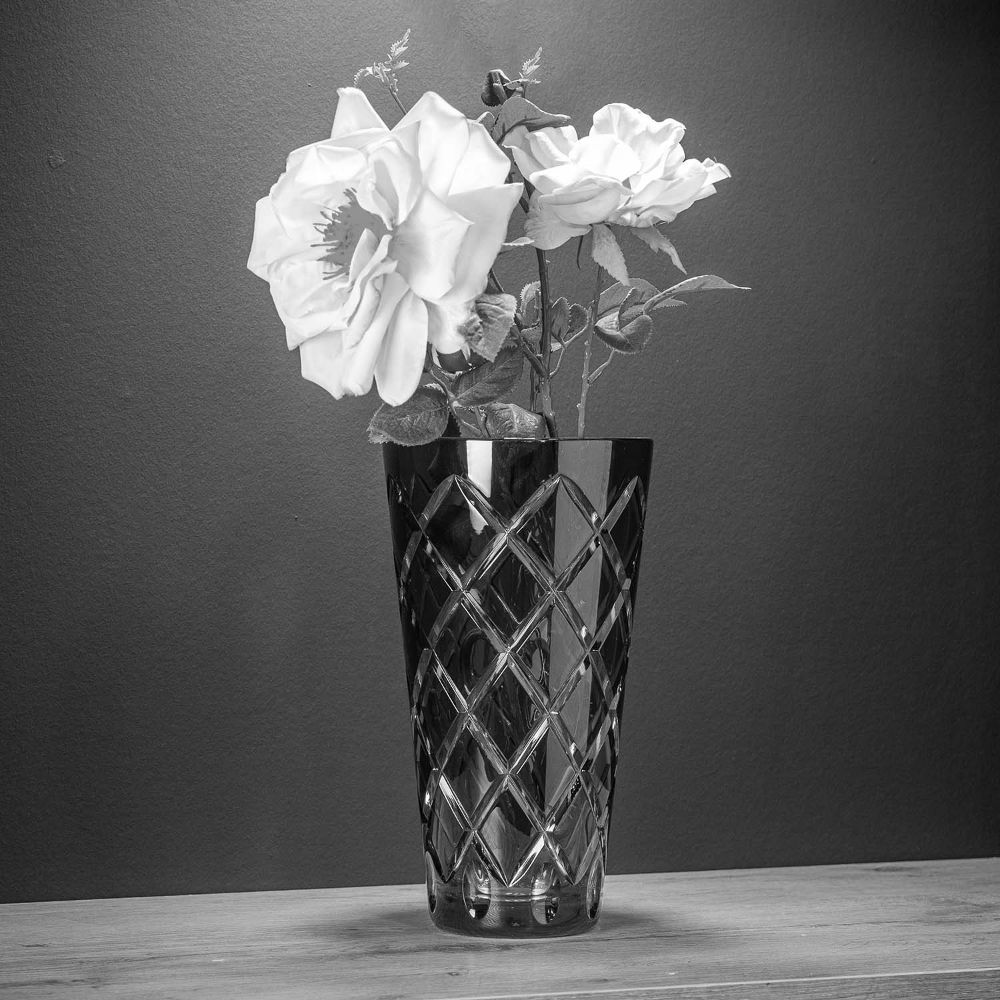 A tall, artfully cut crystal vase with an elegant diamond pattern and a dazzling sheen, containing three bright, fully bloomed roses with leaves. Hand-cut in the style of the Sunline collection, on wood against a dark background.