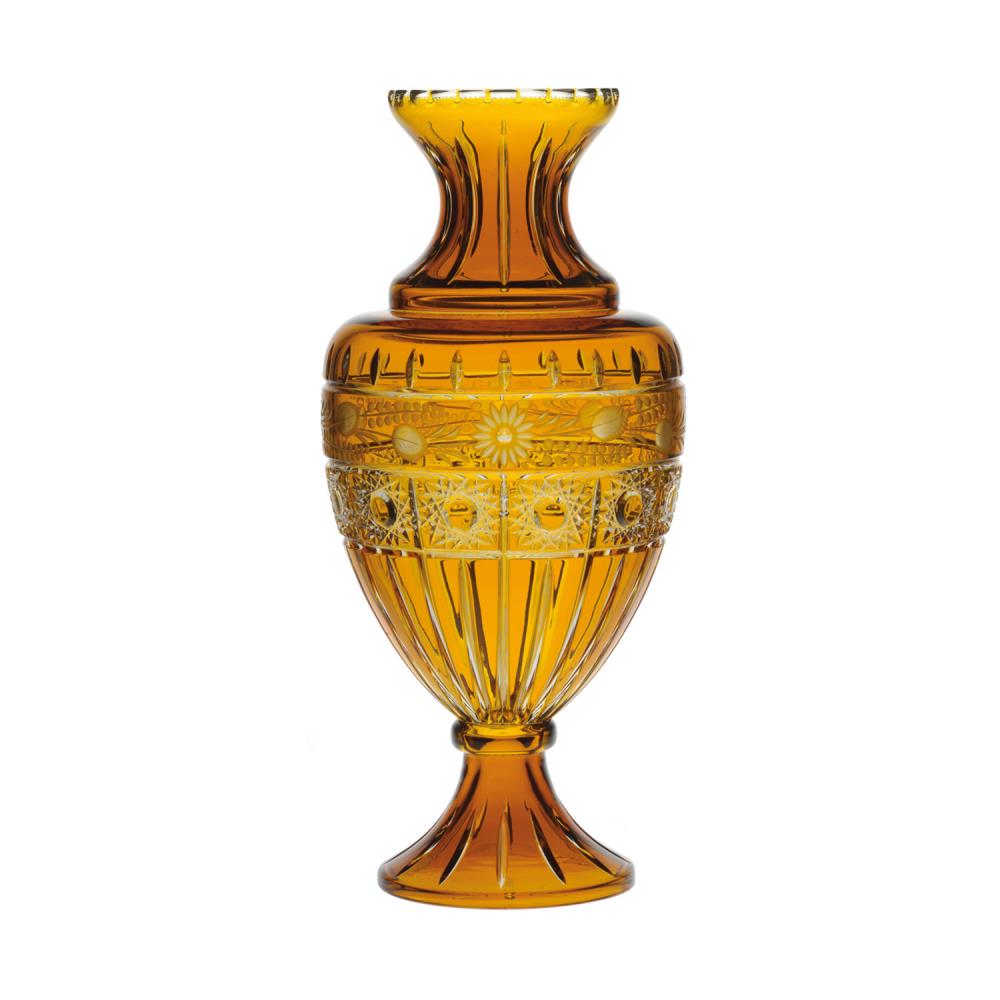 A tall, amber-coloured crystal vase with an elegant silhouette: a narrow neck and a bulbous body. An intricately cut pattern of stylised flowers and geometric shapes. A shiny, light-reflecting surface.