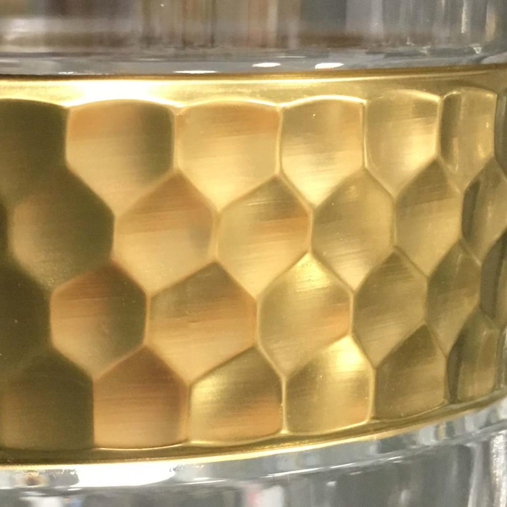 Close-up of a golden, hammered decorative band on a crystal vase. It features a uniform, honeycomb-like hexagonal pattern that reflects light and shimmers luxuriously. Above and below: clear crystal glass.