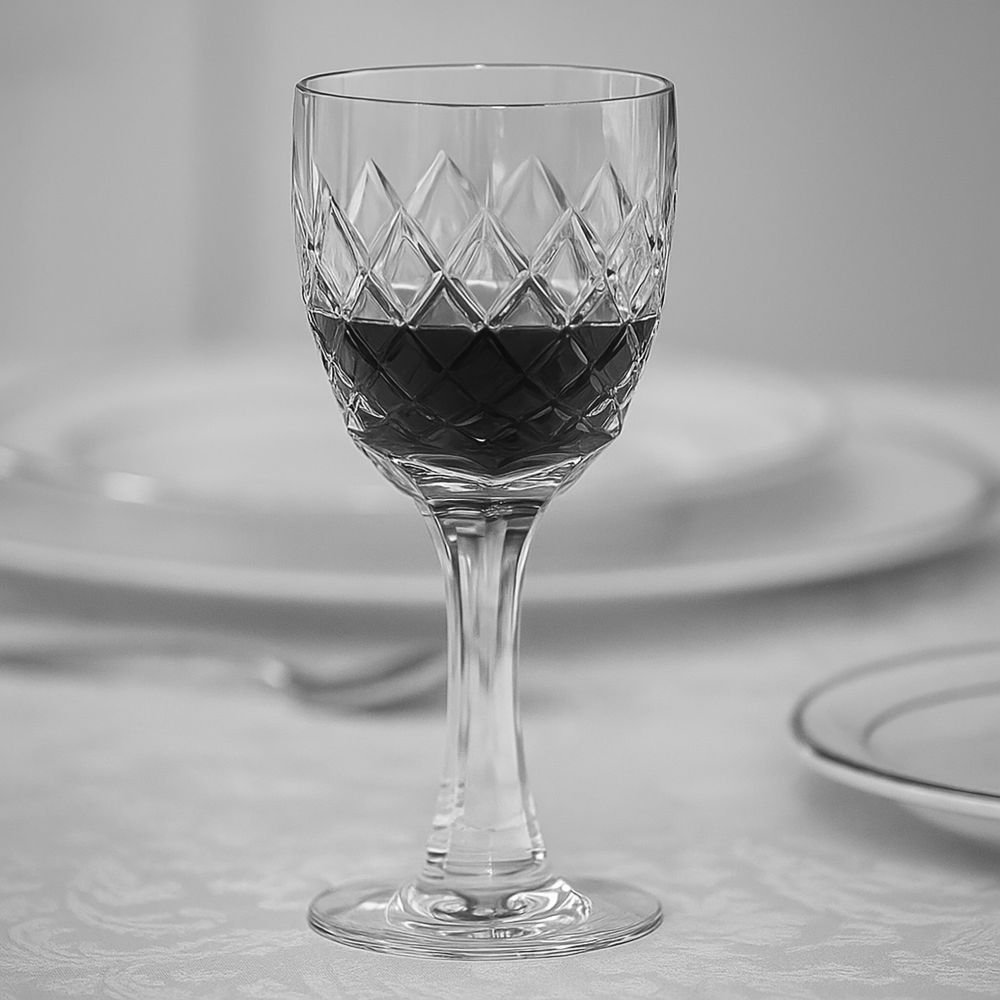 An elegant Venice-style crystal wine glass with a diamond cut sits on a stylishly laid table. It is half-filled with dark red wine and reflects the light. In the blurred background: white plates.