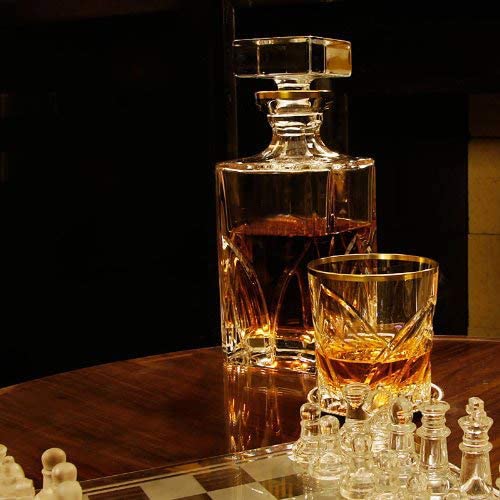 An elegant ‘ARNSTADT KRISTALL’ whisky glass with gold rim and matching cut-glass decanter, both filled with amber whisky, on a polished wooden table. The opulent ‘Wings’ pattern glistens in subdued light. Light chess pieces foreground.